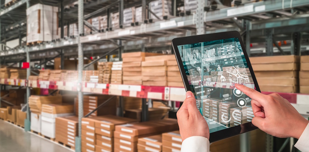 An operator using a digital interface on a tablet to manage warehouse logistics and 3D bin packing optimization. The tablet screen displays real-time AI data overlays, analytics, and spatial maps, with rows of pallet racking and stacked inventory boxes blurred in the background.