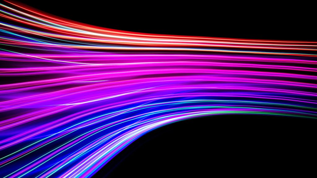 An abstract long-exposure photograph of vibrant, flowing streaks of light in shades of pink, purple, blue, red, and green, curving across a solid black background to create a sense of speed and energy.