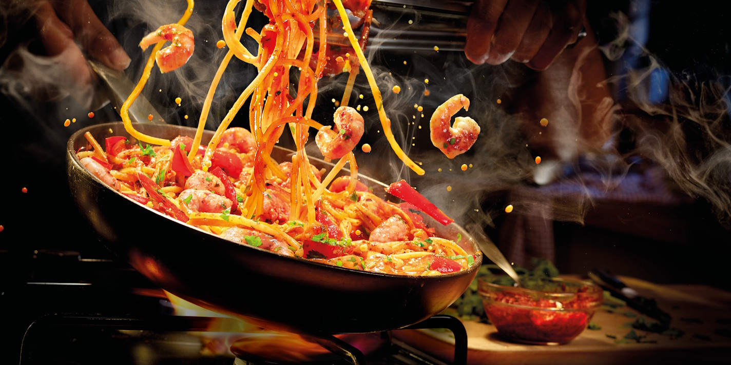 A chef tossess a pan of steaming linguine with prawns and red peppers. Long strands of pasta and several prawns are captured mid-air, surrounded by wisps of white steam and golden droplets of sauce against a dark, warm kitchen background.