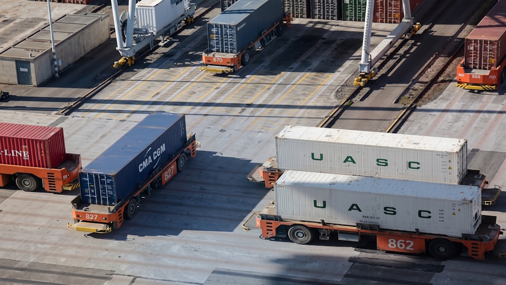 An overhead view of an industrial shipping terminal showing multiple automated guided vehicles (AGVs) carrying large intermodal cargo containers across a paved transit yard, illustrating complex logistics fleet management and routing constraints.