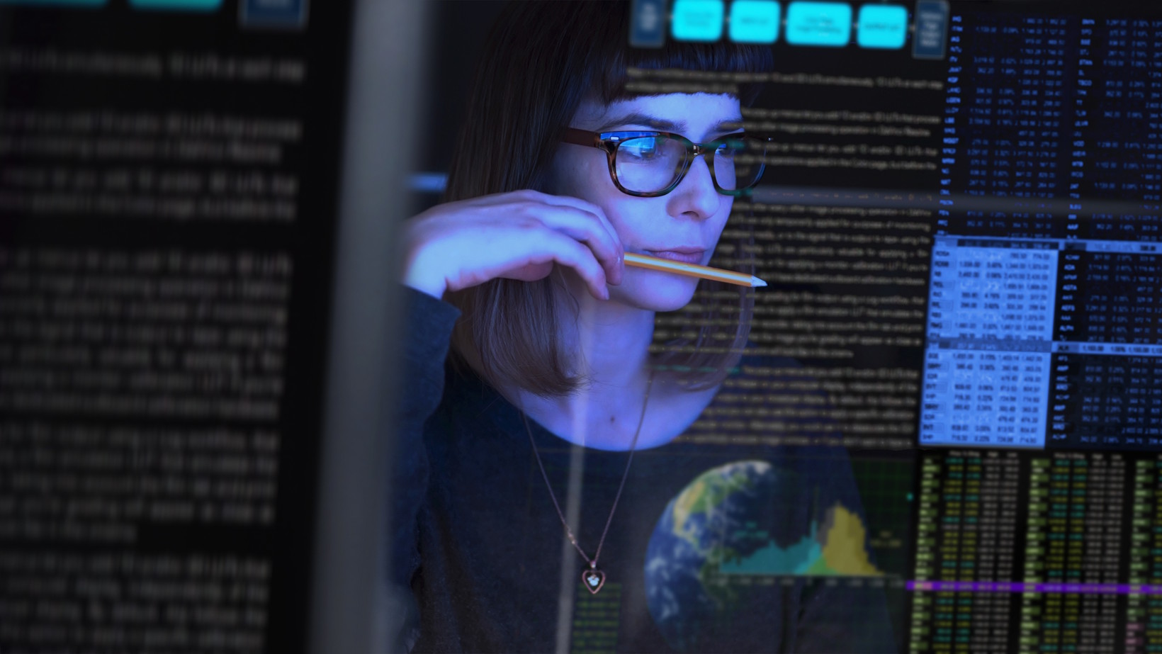 Close-up of a woman with short brown hair and glasses holding a pencil against her chin in deep concentration. She is looking at transparent digital displays filled with lines of code, data tables, and a graphic of Earth, lit by the cool blue glow of the monitors.